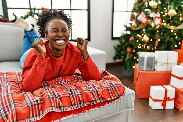 若いアフリカ系アメリカ人女性が腕を上げ 勝利の笑顔を祝う目を閉じて成功に興奮クリスマスツリーでソファの上に横たわっていた 勝者のコンセプト — ストック写真