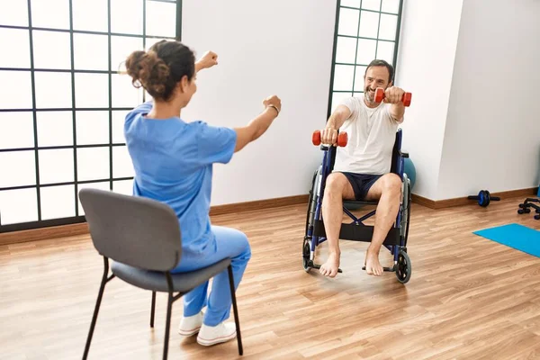 理学療法クリニックで車椅子に座るダンベルを使ったリハビリセッションを持つ中年男性と女性 — ストック写真