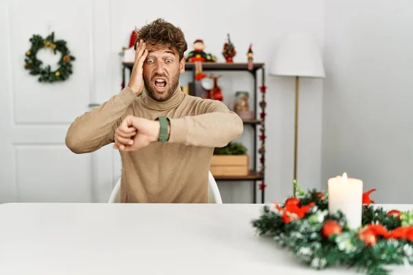 若いですハンサムな男でひげがテーブルの上に座ってクリスマスの装飾を見ながら時計の時間を心配し 遅くなるのを恐れて — ストック写真