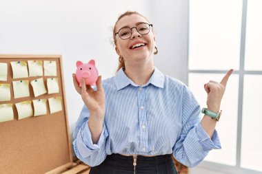 Genç kızıl saçlı kadın domuz kumbarasını elinde tutuyor gülümsüyor mutlu bir şekilde elini ve parmağını yan tarafa gösteriyor. 