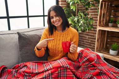 Kanepede oturan genç İspanyol kadın neşeli bir kahve içiyor. Yüzü gülüyor ve mutlu ve doğal bir ifadeyle elini ve parmağını kaldırıyor. 