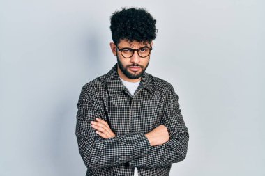Sakallı, gözlüklü, kolları çapraz hareket eden, şüpheci ve gergin genç bir Arap, sorun yüzünden surat asıyor. negatif kişi. 