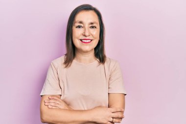 Orta yaşlı İspanyol kadın gündelik kıyafetler giyip kollarını kavuşturup kameraya bakarken gülümsüyor. pozitif kişi. 