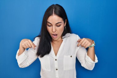 Mavi arka planda dikilen genç İspanyol kadın parmaklarını göstererek reklamını yapıyor, şaşırmış yüz ve ağzını açıyor. 