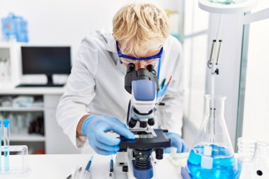 Genç beyaz adam laboratuarda mikroskop kullanarak bilim adamı üniforması giyiyor.