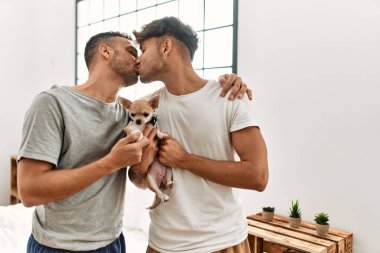 İki İspanyol erkek öpüşüyor ve sarılıyorlar. Yatak odasında chihuahua ile duruyorlar.