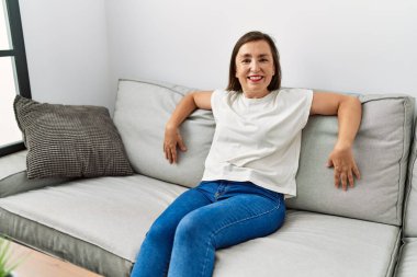 Orta yaşlı İspanyol kadın evdeki kanepede oturmuş gülümsüyor.