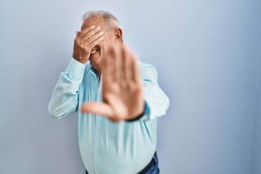 Mavi arka planda gri saçlı yaşlı adam elleriyle gözlerini kapatıyor ve üzgün ve korku ifadesiyle dur hareketi yapıyor. utangaç ve olumsuz kavram. 