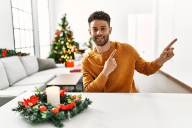 Arap genç adam Noel ağacının yanında masada oturuyor gülümsüyor ve kameraya bakıyor iki eliyle ve parmaklarıyla kenarı işaret ediyor.. 