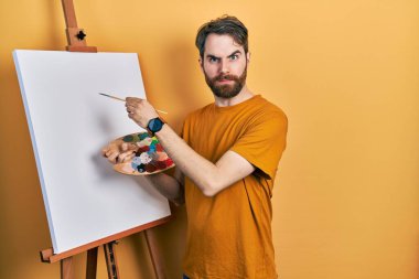 Beyaz, sakallı, palet çizen beyaz bir adam. Resim sehpası kuşkucu ve gergin duruyor. Problemden dolayı surat asıyor. negatif kişi. 