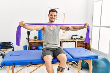 Genç İspanyol adam klinikteki masaj masasında oturan elastik bant kullanarak kolunu esnetiyor.