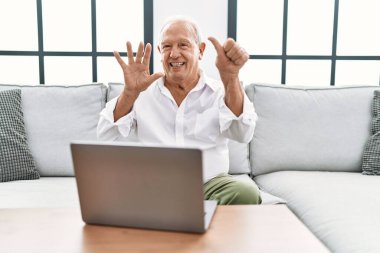 Üst düzey bir adam evinde dizüstü bilgisayar kullanıyor. Koltukta oturuyor ve 6 numaralı parmaklarıyla işaret ediyor. Kendinden emin ve mutlu bir şekilde gülümsüyor.. 