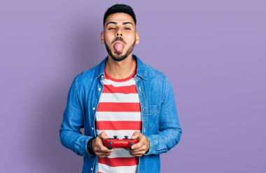 Young hispanic man with beard playing video game holding controller sticking tongue out happy with funny expression. 