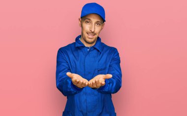 Kel, sakallı, işçi tulumu giyen, elleri avuçlarında gülümseyen ve el hareketi yapan bir adam. Bekleme ve koruma 