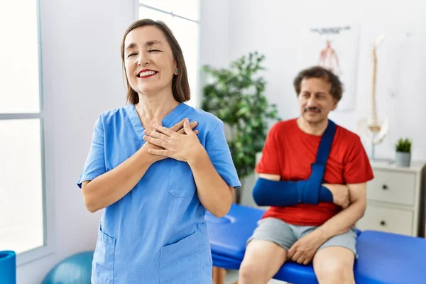 Middle age doctor woman with patient with arm injury at rehabilitation clinic smiling with hands on chest, eyes closed with grateful gesture on face. health concept.