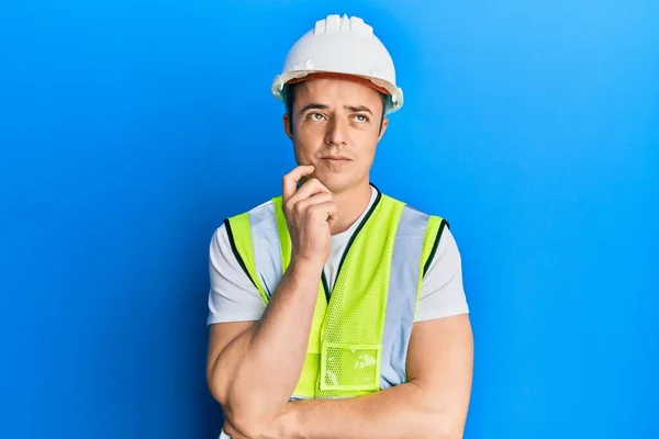 Handsome young man wearing safety helmet and reflective jacket serious face thinking about question with hand on chin, thoughtful about confusing idea
