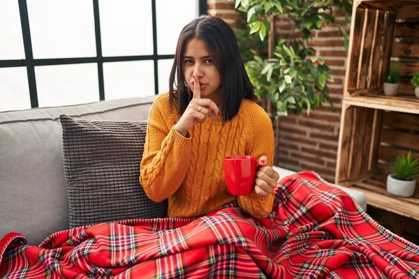 Genç İspanyol kadın kanepede oturup kahvesini yudumluyor ve parmakları dudaklarında sessiz olmak istiyor. sessizlik ve gizli konsept. 