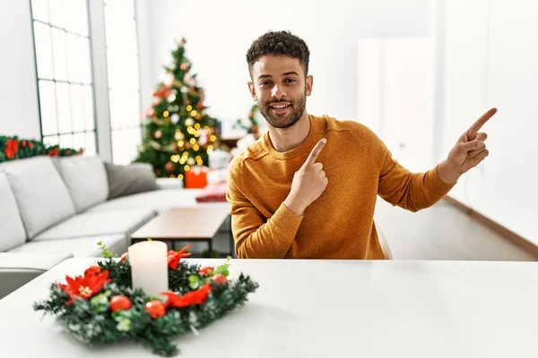 Arap genç adam Noel ağacının yanında masada oturuyor gülümsüyor ve kameraya bakıyor iki eliyle ve parmaklarıyla kenarı işaret ediyor.. 