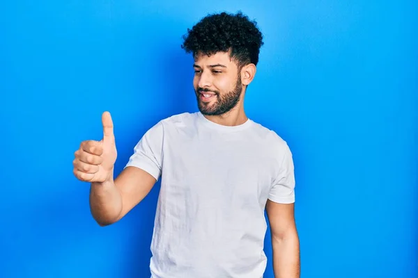 髭を生やした若いアラブ人男性は 誇りに思っているように見えるカジュアルな白いTシャツを着て 親指を横にジェスチャーアップ笑顔 — ストック写真