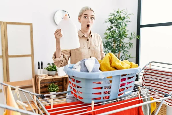 Young Caucasian Woman Doing Laundry Holding Detergent Bottle Scared Amazed — стоковое фото