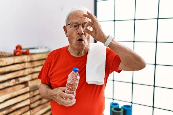 Senior man wearing sportswear and towel at the gym doing ok gesture shocked with surprised face, eye looking through fingers. unbelieving expression.