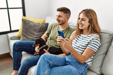 Genç İspanyol çift mutlu bir şekilde gülümsüyor. Akıllı telefon kullanıyor. Kanepede köpeğiyle oturuyor..