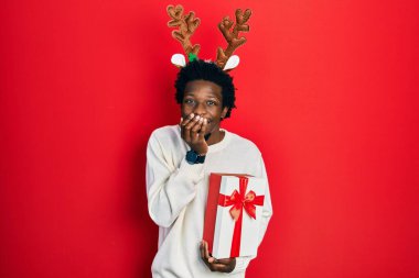 Young african american man wearing deer christmas hat holding gift laughing and embarrassed giggle covering mouth with hands, gossip and scandal concept 