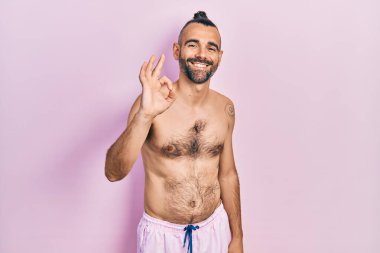Young hispanic man shirtless wearing swimsuit smiling positive doing ok sign with hand and fingers. successful expression. 