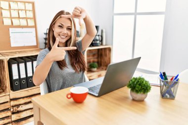 Genç esmer kadın ofiste dizüstü bilgisayarla çalışıyor gülümsüyor ve mutlu yüzlü eller ve parmaklarla çerçeve yapıyor. yaratıcılık ve fotoğrafçılık kavramı. 