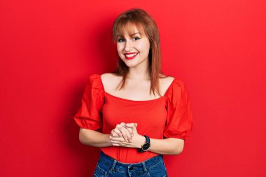 Kızıl saçlı genç bir kadın elleri bağlı, günlük kırmızı tişörtler giyiyor ve mutlu ve rahat bir şekilde gülümsüyor. Başarı ve iyimserlik 