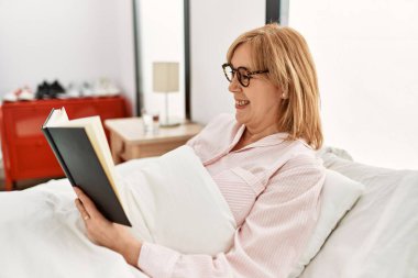 Orta yaşlı sarışın kadın evde yatağında kitap okuyor..