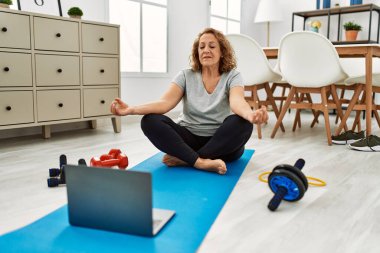 Orta yaşlı beyaz bir kadın evde laptopla yoga eğitimi veriyor.