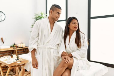 Havlu giymiş genç Latin bir çift, güzellik merkezinde durup yan tarafa bakıyor, rahat bir görünüm takınıyor ve kendine güvenen bir gülümseme takınıyor.. 