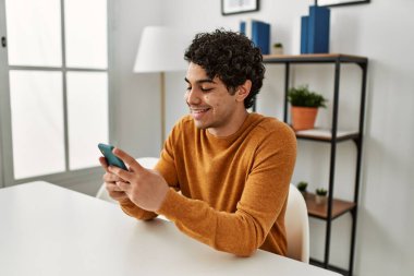 Genç İspanyol adam akıllı telefon kullanıyor. Evdeki masada oturuyor..