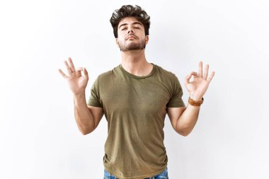 İzole edilmiş beyaz arka planda duran İspanyol adam rahatlamış ve parmaklarıyla meditasyon hareketi yaparken gözleri kapalı gülümsüyor. yoga konsepti. 