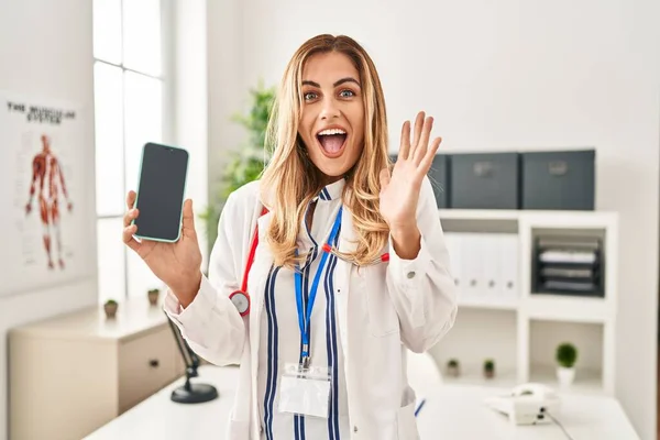 Young Blonde Doctor Woman Working Clinic Showing Smartphone Screen Celebrating — Stock Photo, Image