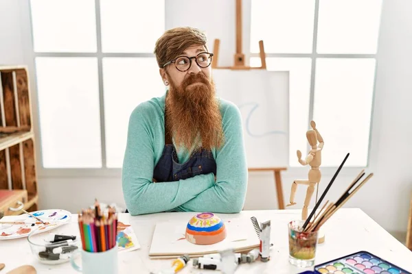 Redhead man with long beard painting clay bowl at art studio smiling looking to the side and staring away thinking.