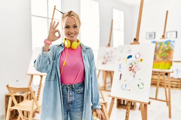 Young caucasian girl at art studio smiling positive doing ok sign with hand and fingers. successful expression.