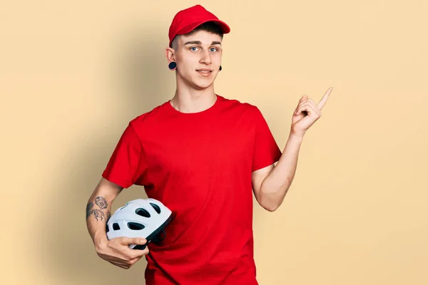 Young caucasian boy with ears dilation wearing delivery uniform and cap holding bike helmet smiling happy pointing with hand and finger to the side