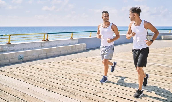 İki İspanyol erkek, sportif, gülümseyen bir çift, deniz kenarında koşuyorlar.