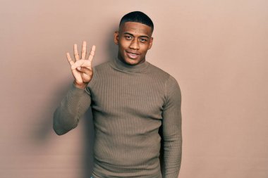Balıkçı yaka kazak giyen genç siyah bir adam gülümseyerek kendinden emin ve mutlu bir şekilde parmaklarıyla dört numarayı gösteriyor ve işaret ediyor.. 