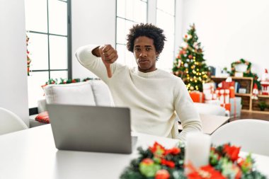 Genç Afro-Amerikalı adam dizüstü bilgisayar kullanıyor. Noel ağacının yanında oturuyor. Mutsuz ve sinirli görünüyor. kötü ifade. 