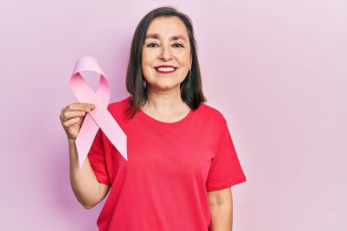 Orta yaşlı İspanyol kadın pembe kanser kurdelesi tutuyor. Pozitif ve mutlu görünüyor. Kendine güvenen bir gülümsemeyle dişlerini gösteriyor. 