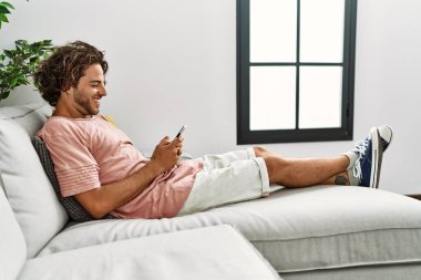Genç İspanyol adam mutlu bir şekilde gülümsüyor. Evdeki kanepede oturmuş akıllı telefon kullanıyor..
