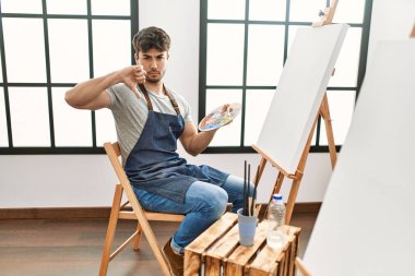Young hispanic man painting at art studio with angry face, negative sign showing dislike with thumbs down, rejection concept 