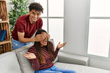 Man surprising his girlfriend with hands on eyes sitting on the sofa at home.