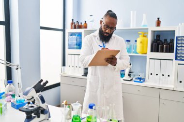 Bilim adamı üniforması giyen genç Afro-Amerikan adam laboratuardaki panoya yazı yazıyor.