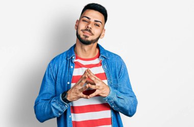 Sakallı, sıradan kot ceket giyen genç İspanyol adam ellerini birleştirip gülümseyerek gevşemiş ve neşeliydi. Başarı ve iyimserlik 