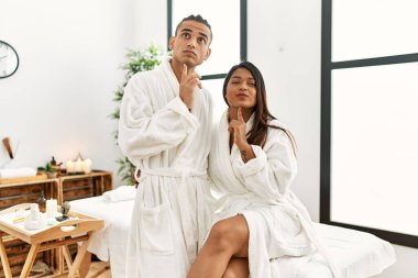 Havlu giymiş genç Latin bir çift güzellik merkezinde durup parmakları çenesinde şüpheye yoğunlaşmışlar ve merak içindeler. 