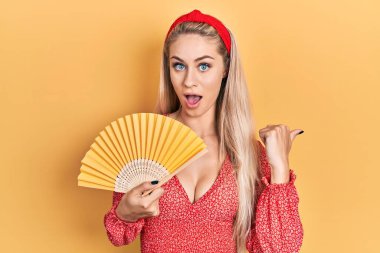 Beyaz bir kadın el sallıyor yazın serinleyen havayı işaret ediyor başparmağını yana uzatıyor ve ağzı açık gülümsüyor. 